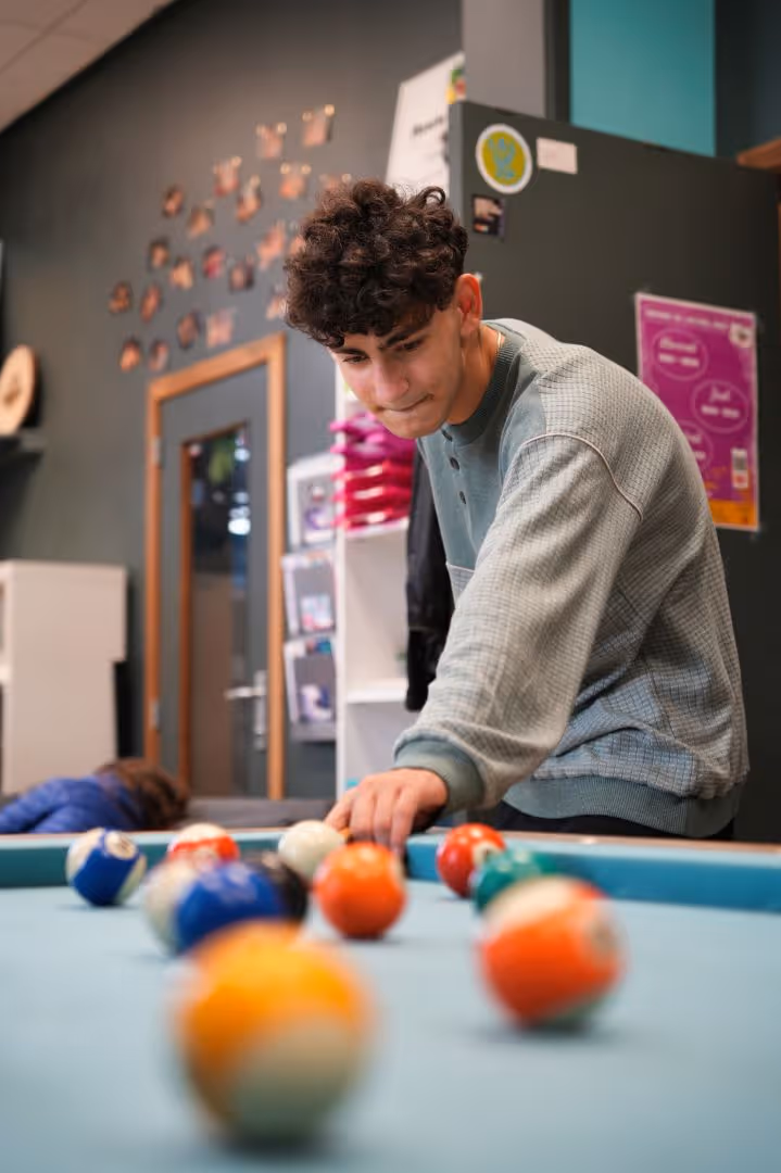 Un jeune homme concentré jouant au billard dans une salle avec un mur décoré de photos.
