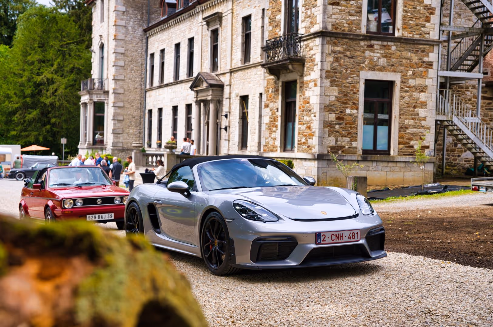 Voiture de sport grise Porsche devant une vieille maison en pierre avec des personnes debout à l'arrière.
