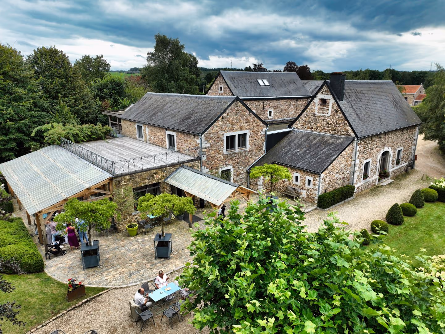 Vue aérienne d'une grande maison en pierre avec terrasse, jardin et personnes assises dehors.