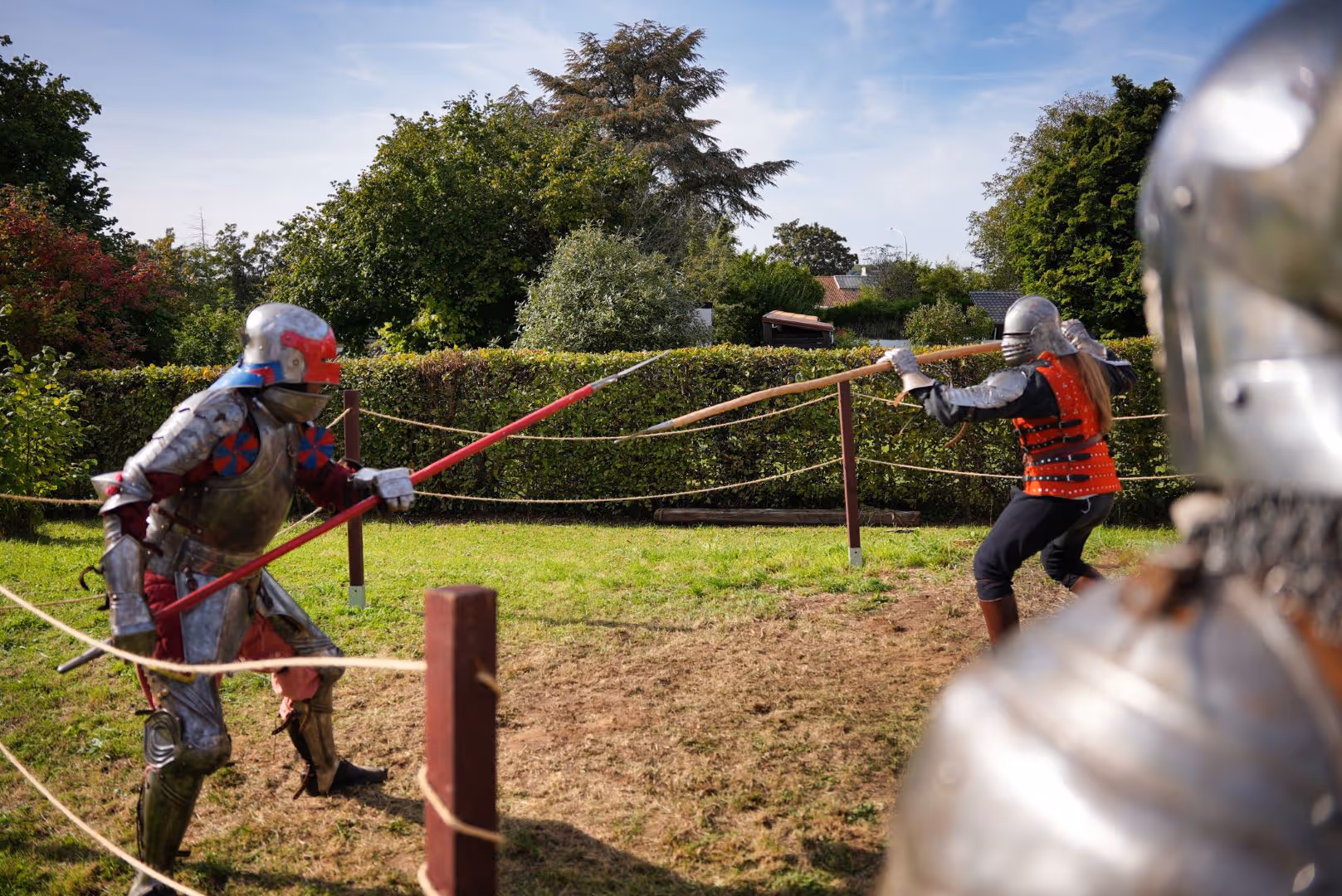 Deux combattants en armure médiévale s'affrontent à la lance sur un terrain gazonné en plein air.