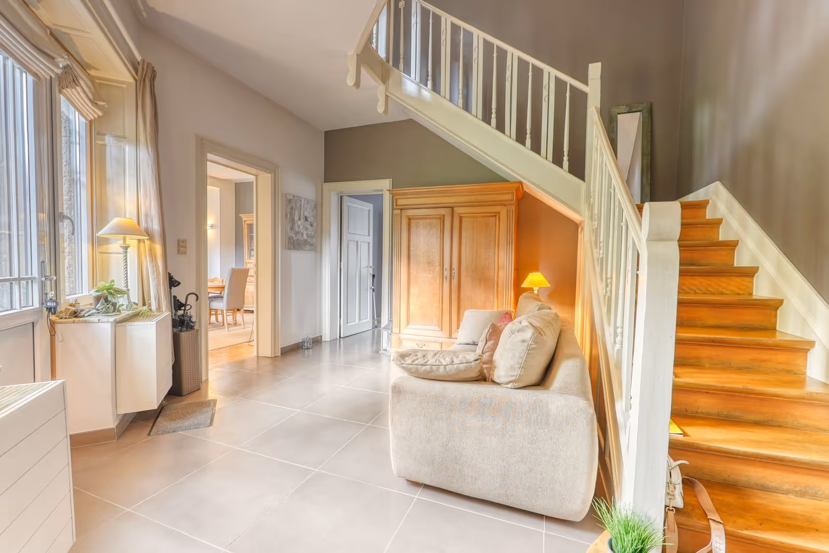 Entrée lumineuse avec canapé beige, armoire, escalier en bois et porte vers une salle à manger.