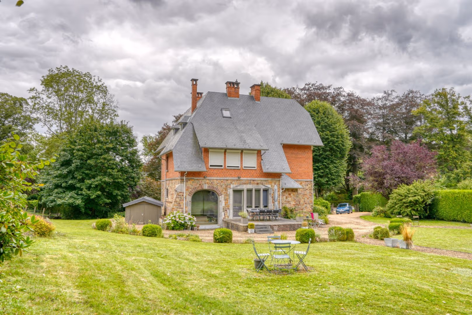 Maison en brique avec toit en ardoise dans un grand jardin vert sous un ciel nuageux.
