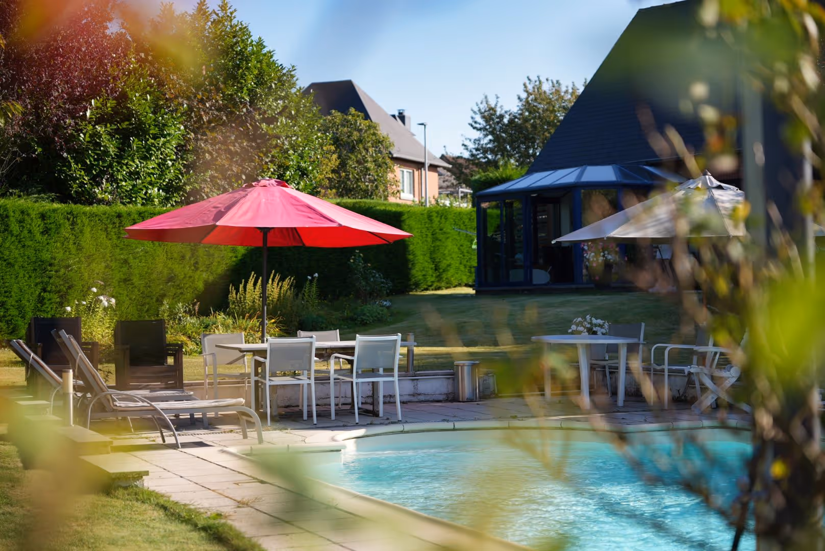 Piscine dans un jardin avec chaises longues, tables, parasols rouge et blanc, et maison en arrière-plan.