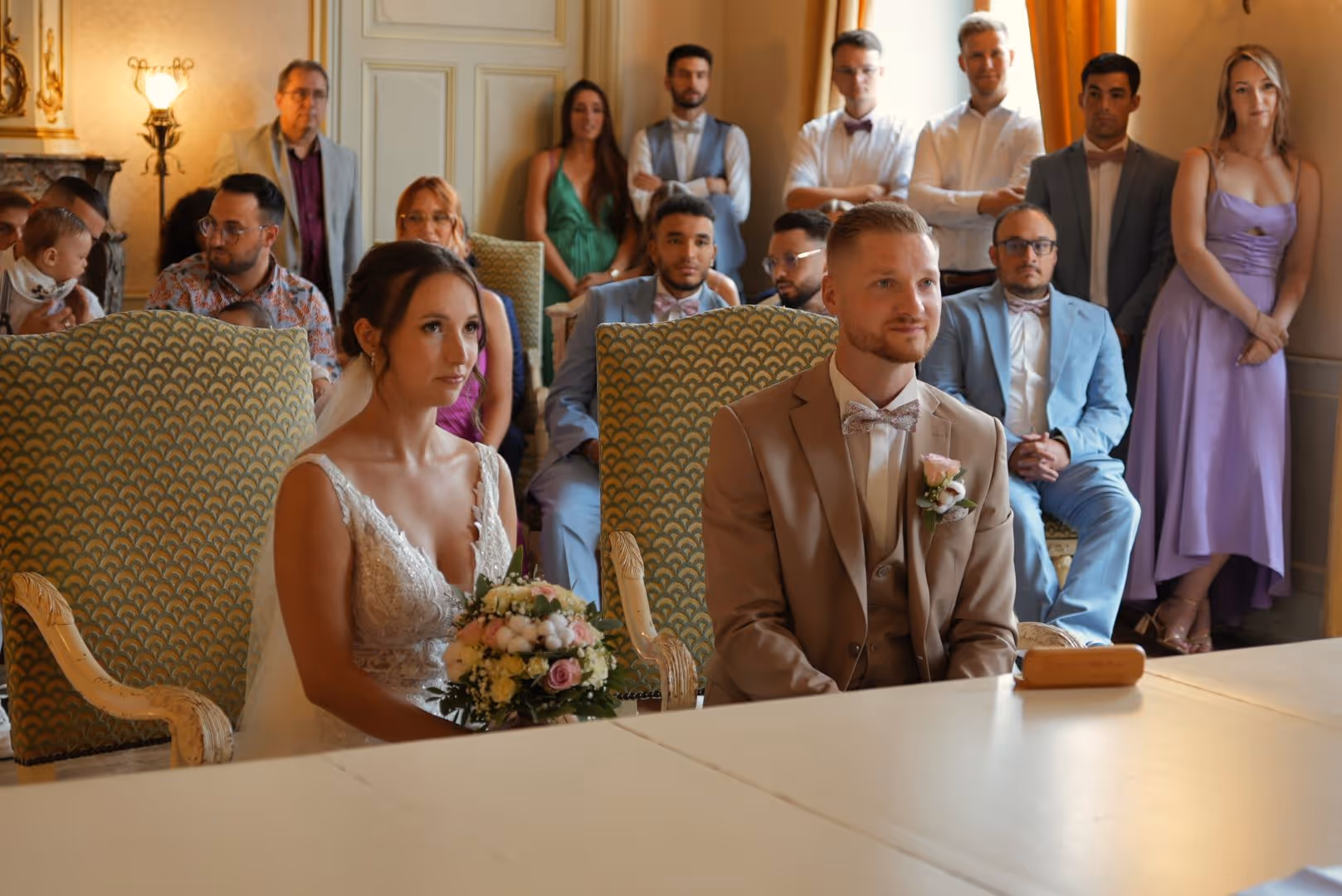 Jeune couple en tenue de mariage assis lors d’une cérémonie civile entouré de leurs invités.