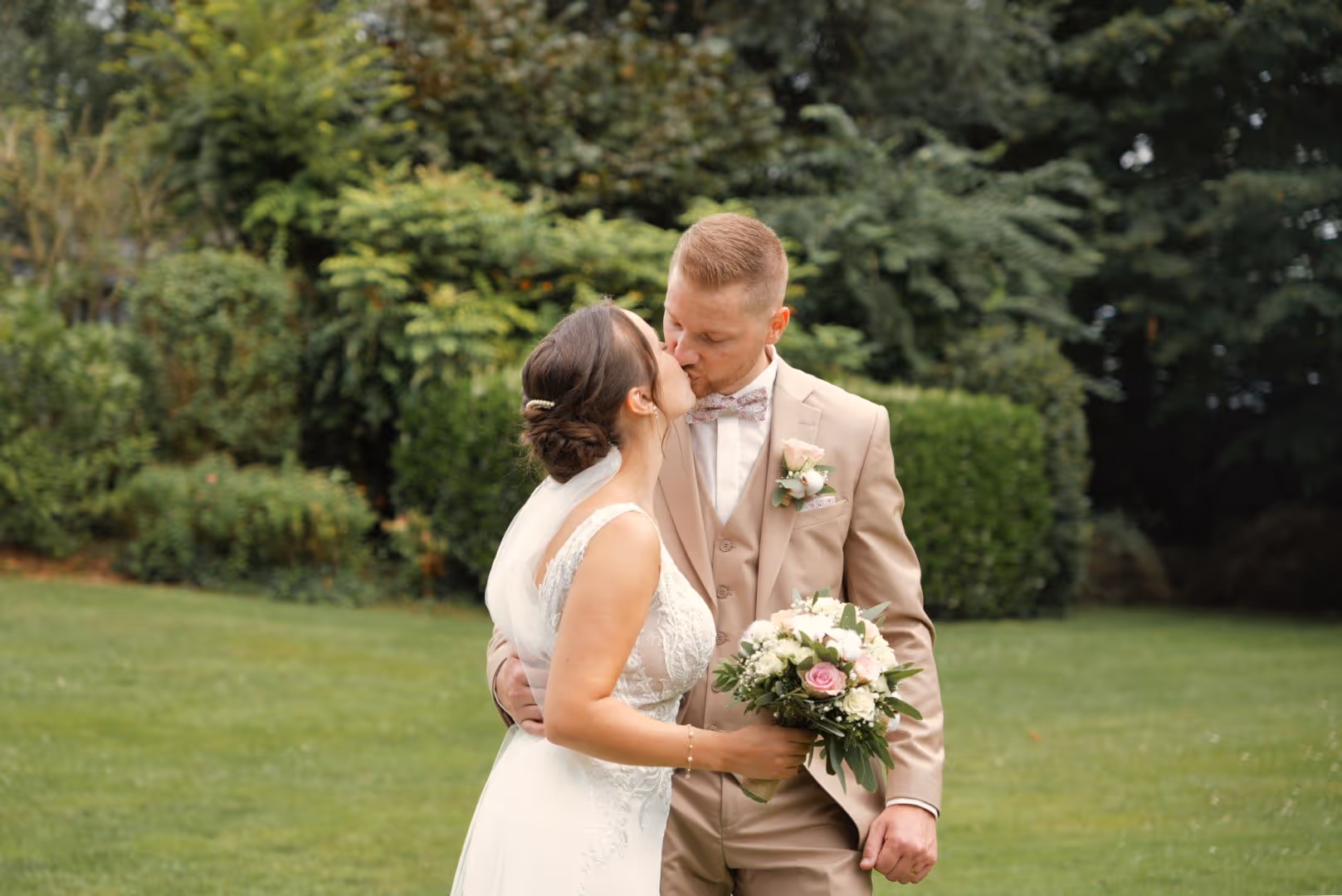 Mariée et marié s'embrassant dans un jardin, la mariée tient un bouquet de fleurs.