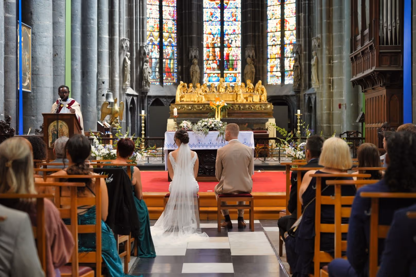 Mariée et marié assis face à l'autel dans une église avec invités et prêtre en arrière-plan.