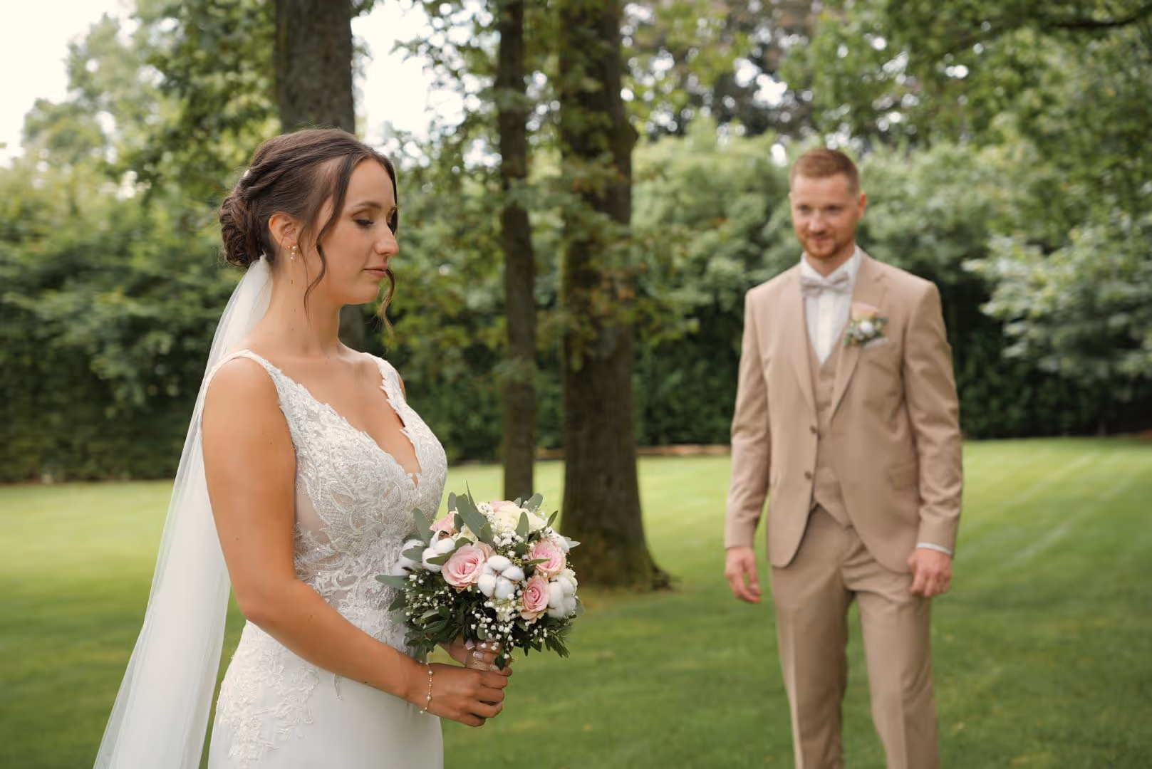 La mariée tient un bouquet fleuri tandis que le marié en costume beige se tient en arrière-plan dans un jardin.