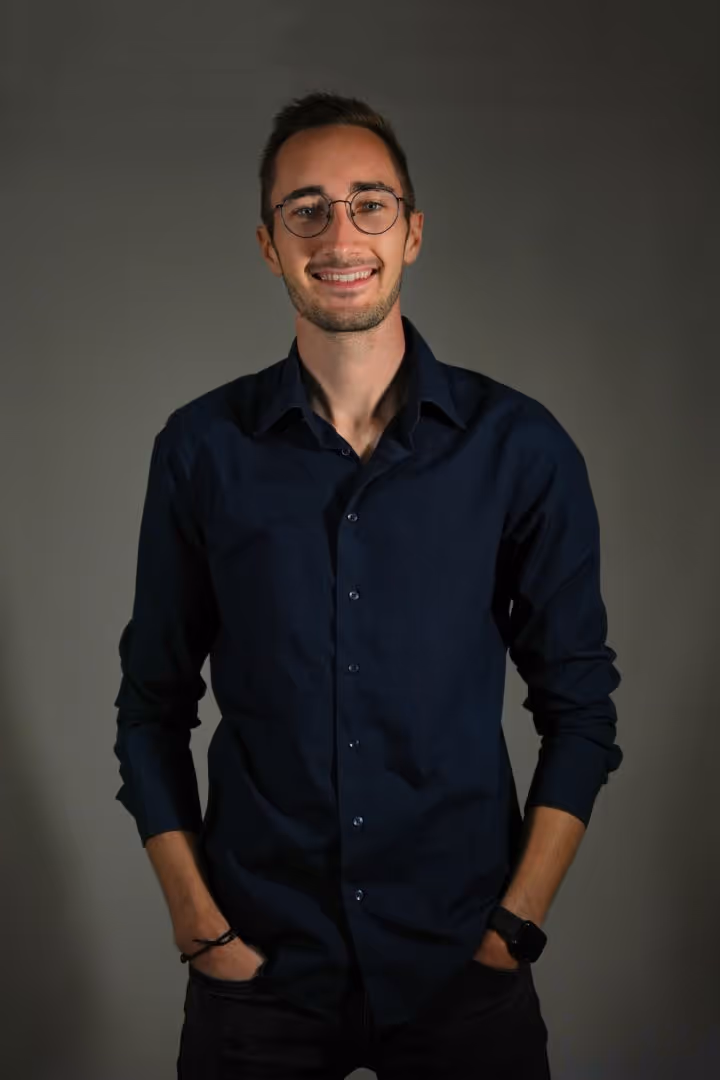 Jeune homme souriant portant une chemise bleue marine et des lunettes, mains dans les poches.