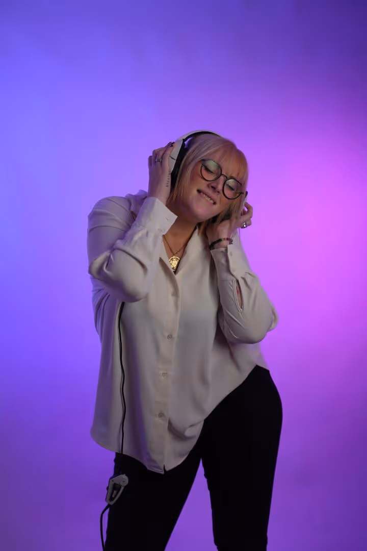 Femme avec lunettes et chemise blanche écoutant de la musique avec un casque sur fond violet.