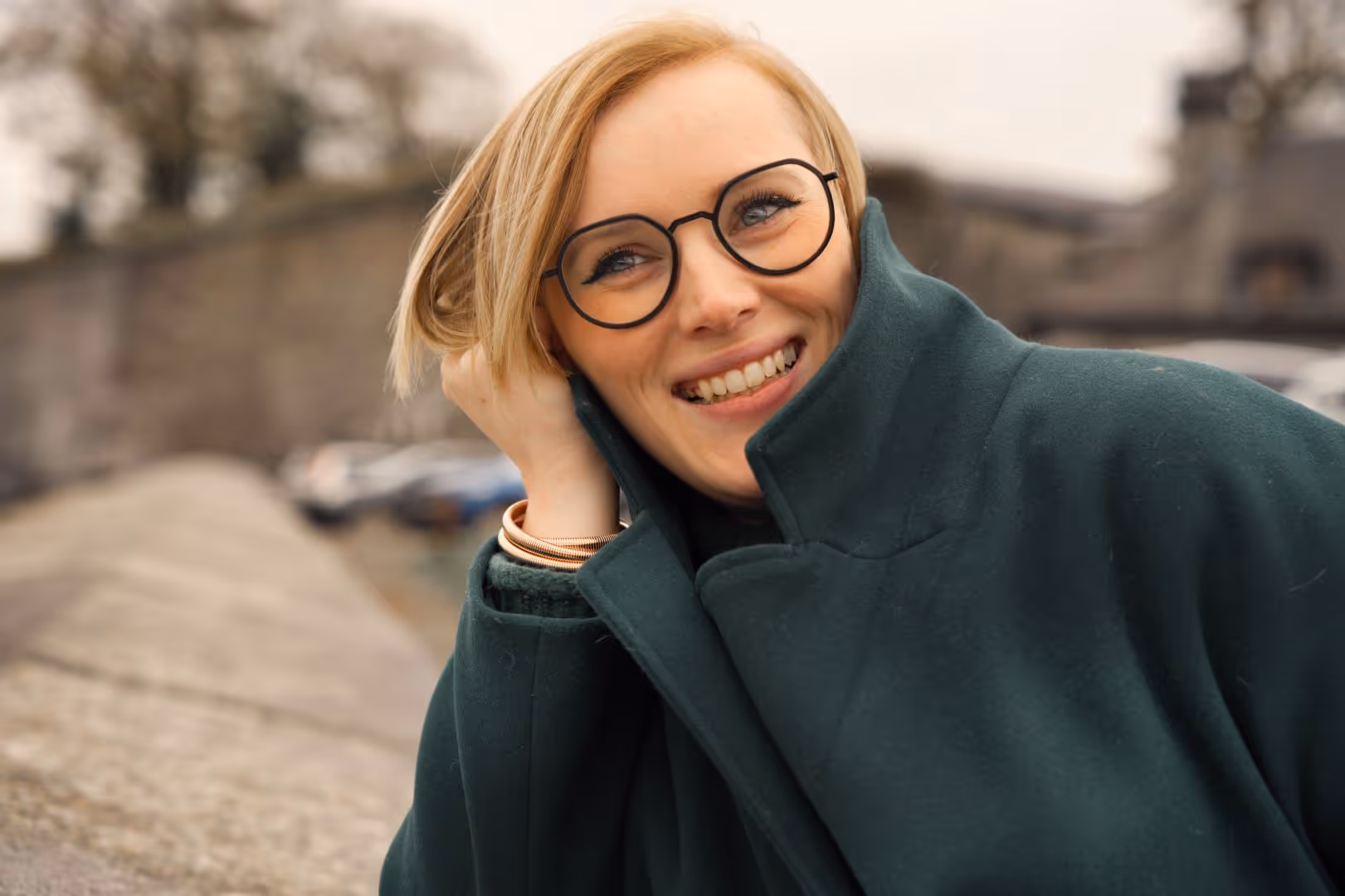 Femme souriante aux cheveux blonds et lunettes rondes portant un manteau vert foncé.