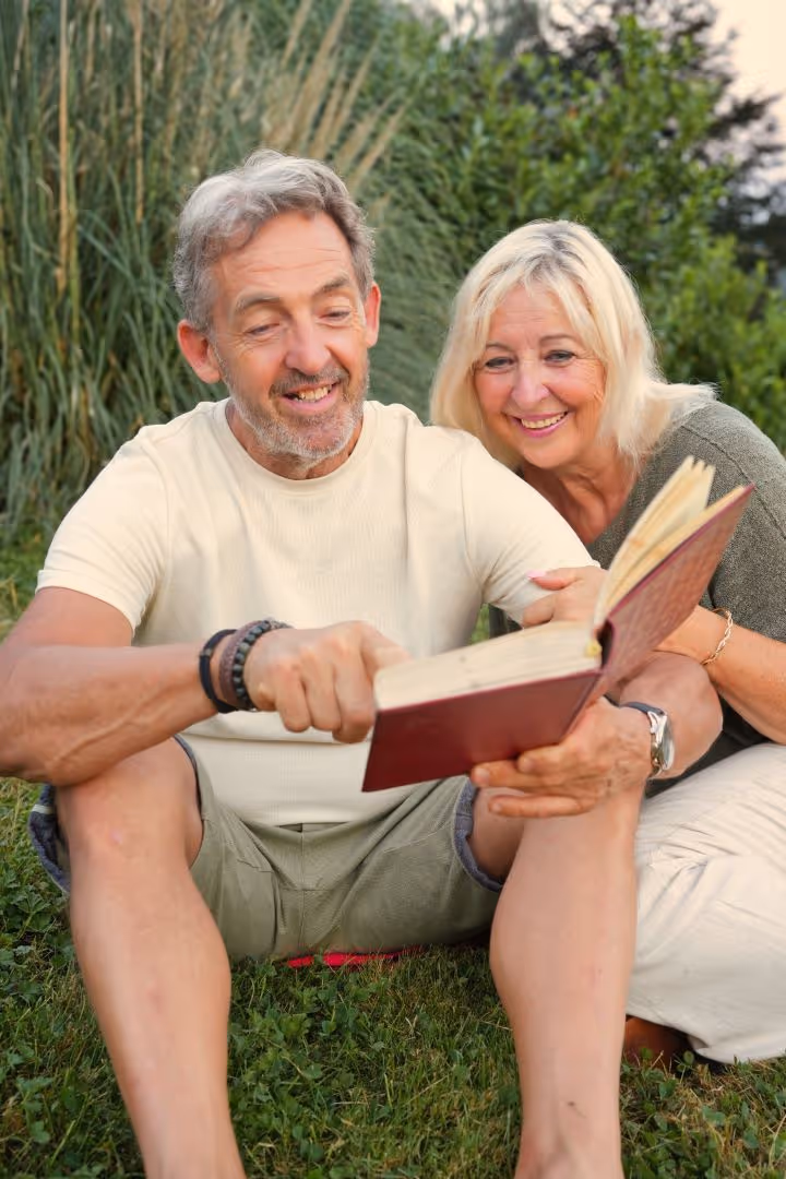 Un homme et une femme mûrs assis dans l'herbe, lisant un livre ensemble avec un sourire.