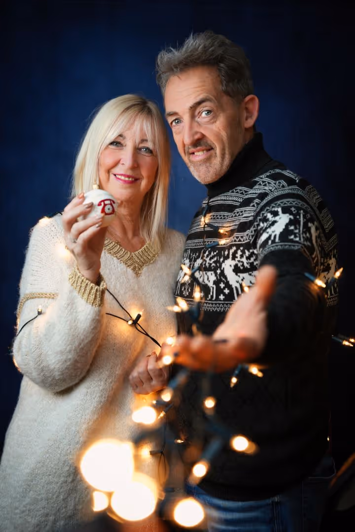 Couple souriant avec pulls de Noël tenant des lumières et une boule de Noël blanche.