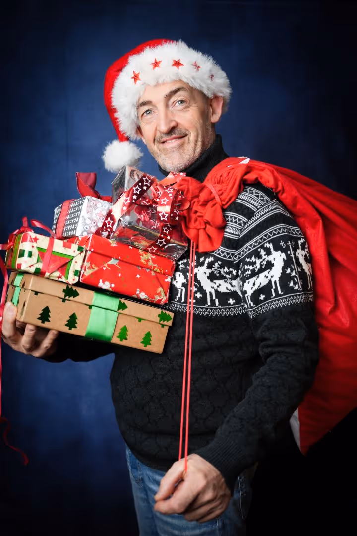 Homme avec un bonnet de Noël tenant plusieurs cadeaux et un sac rouge sur l'épaule.