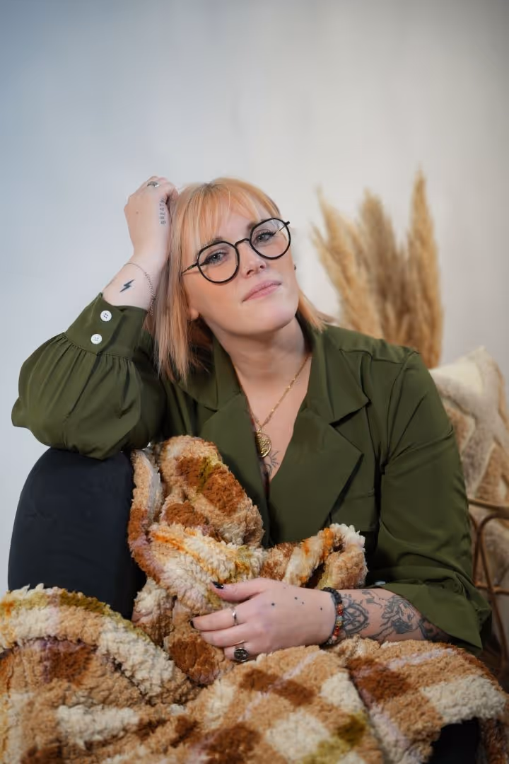 Femme aux cheveux blonds avec lunettes rondes, vêtue d'un manteau vert et tenant une couverture tricolore.