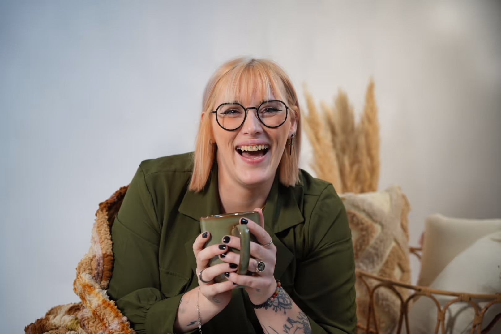 Jeune femme souriante avec lunettes, manteau vert, tenant une tasse verte, assise près d’un fauteuil avec coussins.