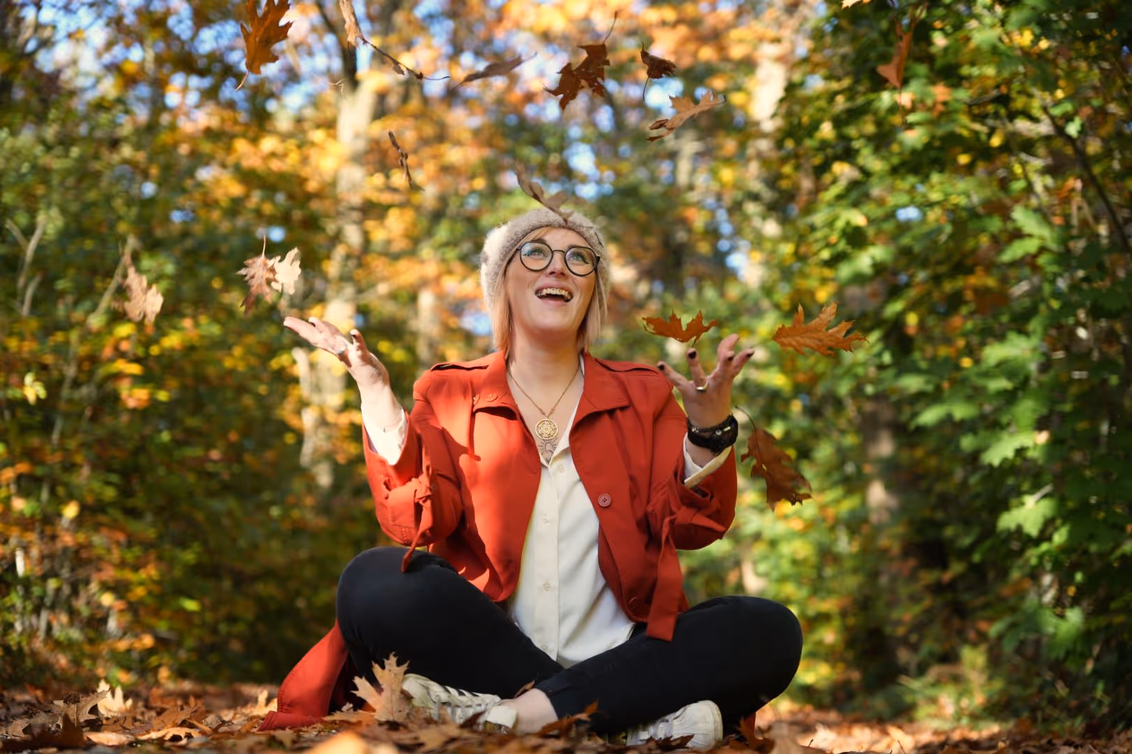 Femme assise en tailleur dans une forêt d’automne, lançant des feuilles avec joie.