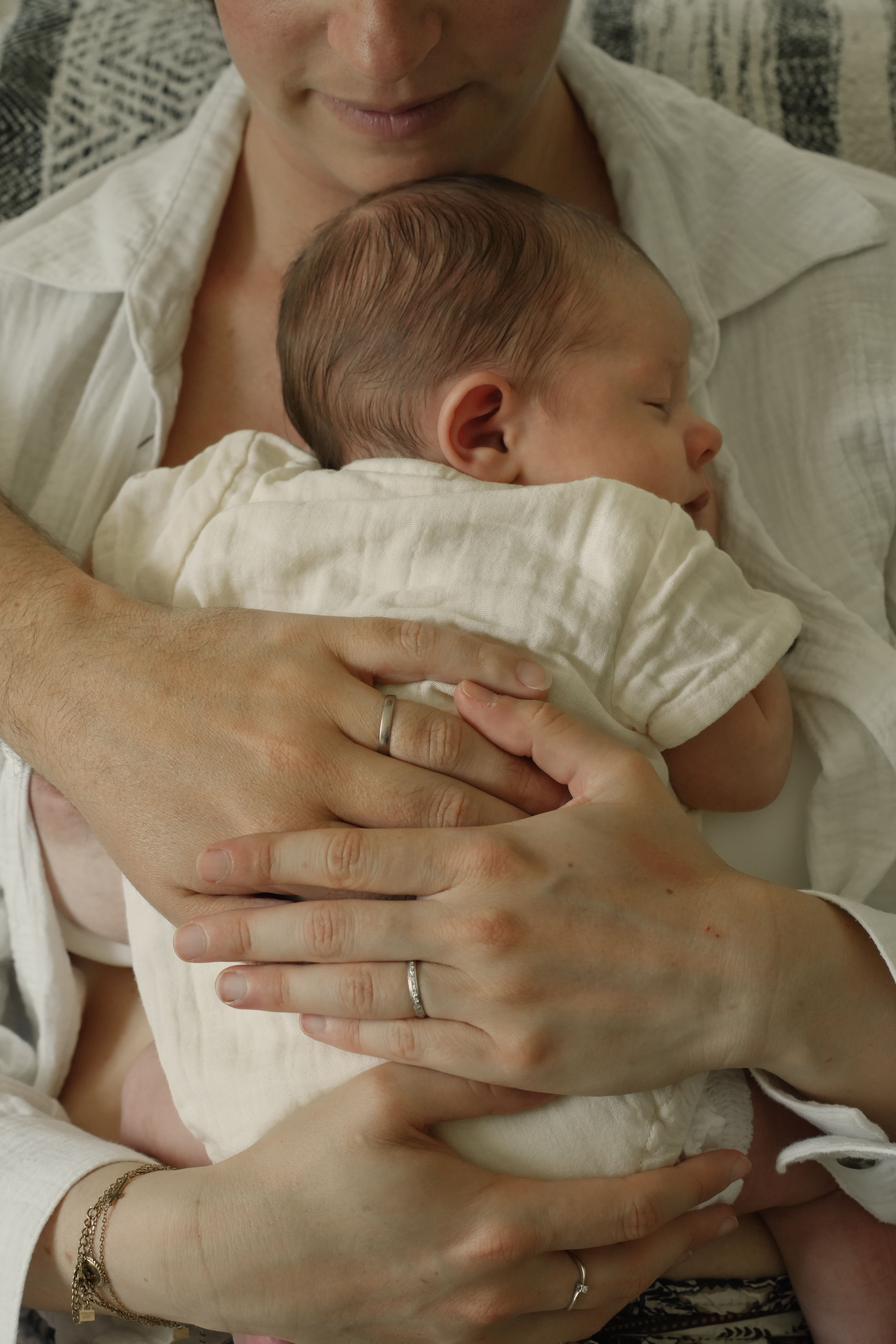 Un bébé endormi dans les bras d'une personne portant une chemise blanche et deux alliances en argent.