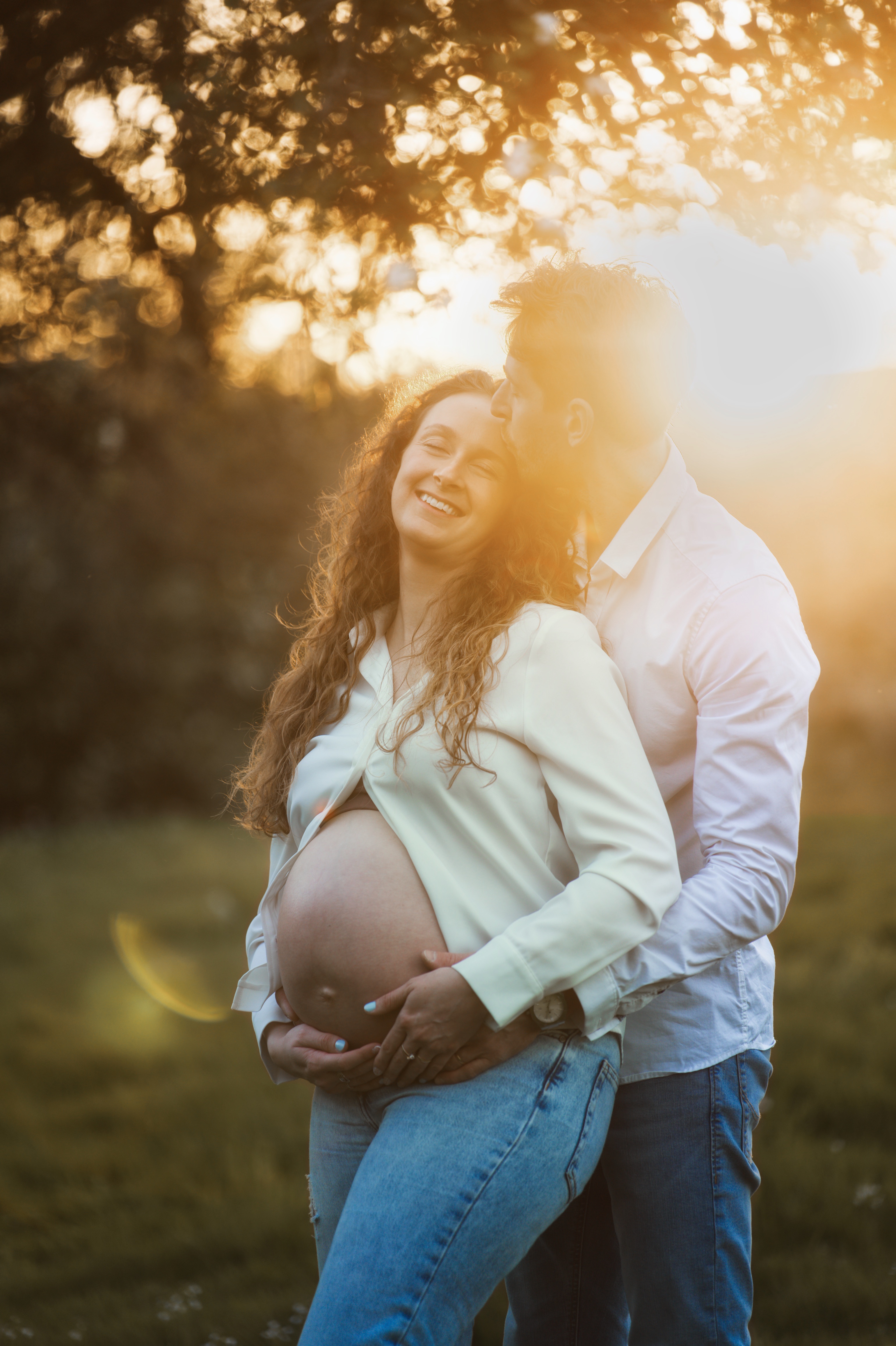 Couple en plein air au soleil, homme embrassant femme enceinte souriante aux cheveux bouclés.