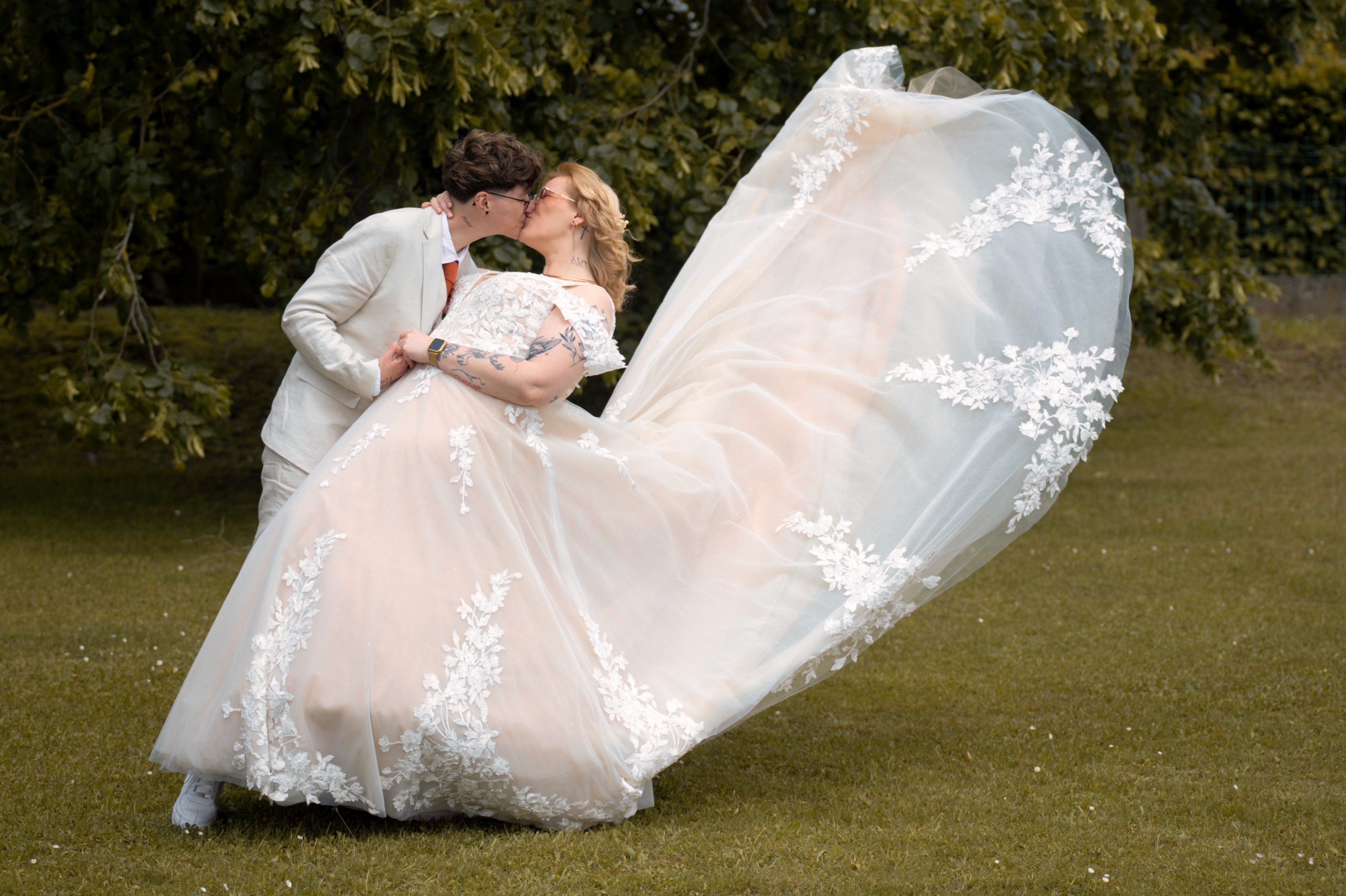 Couple s'embrassant en extérieur, la femme porte une robe de mariée flottante avec dentelle blanche.