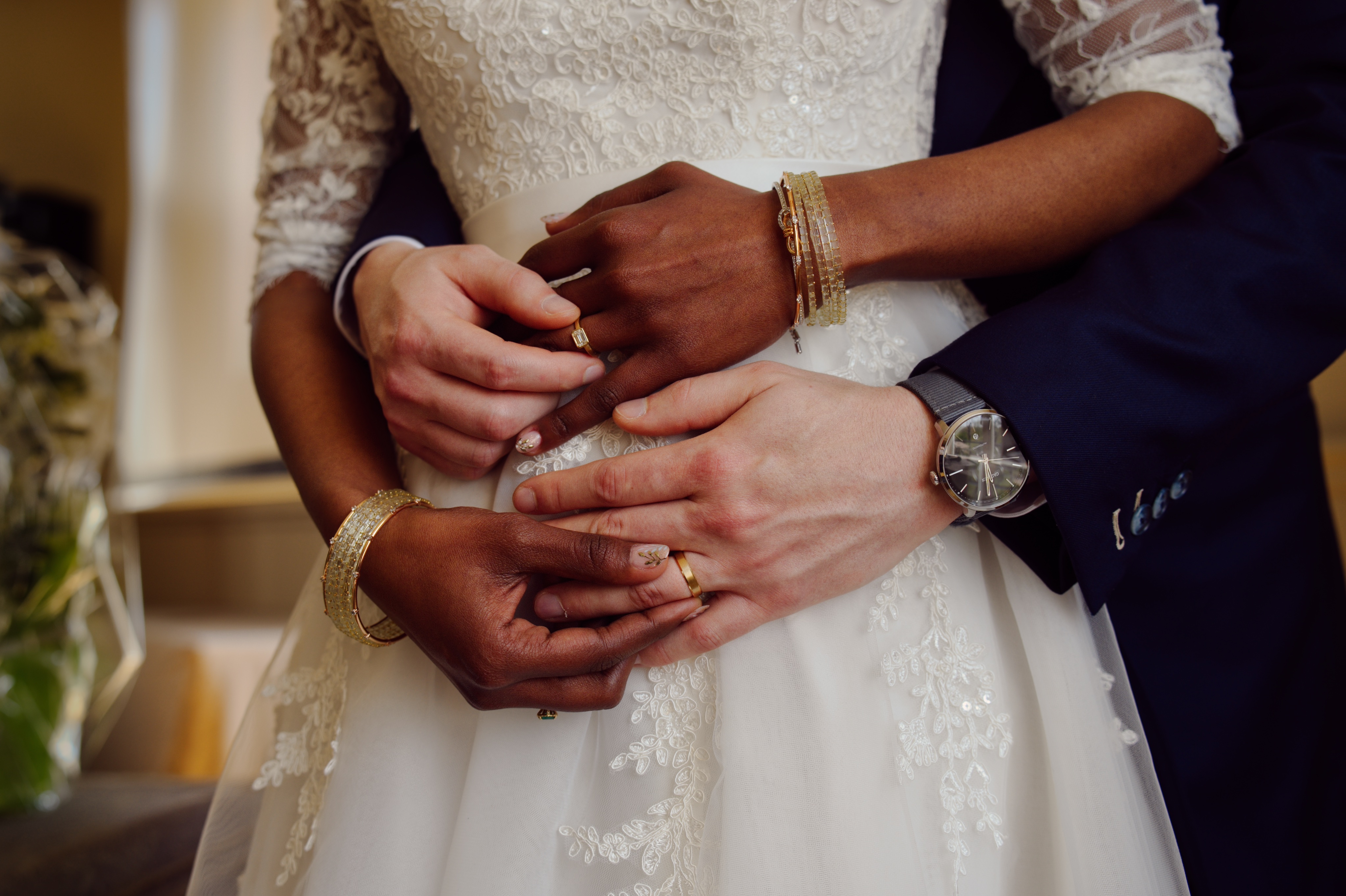 Mains du couple marié entrelacées, elle en robe blanche avec bracelets dorés, lui en costume bleu.