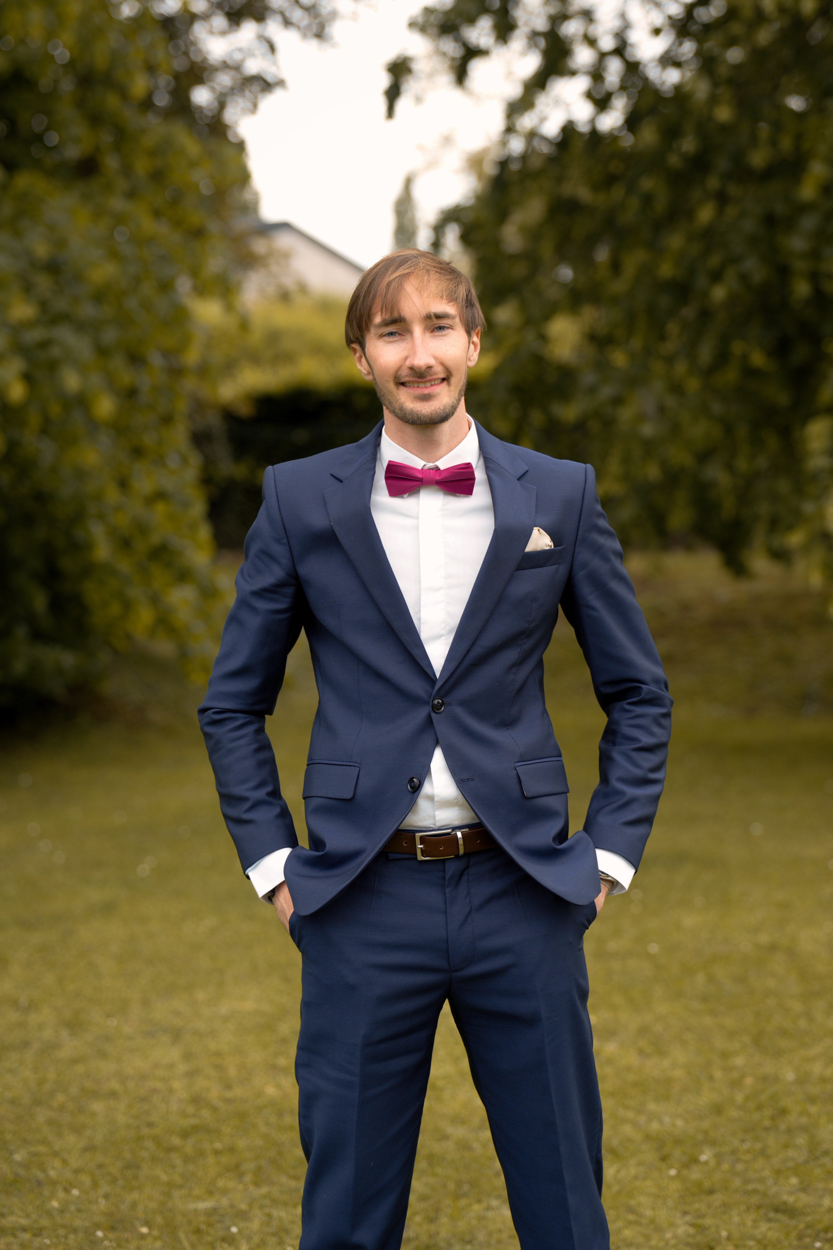 Jeune homme souriant en costume bleu marine, chemise blanche et nœud papillon rouge en plein air.
