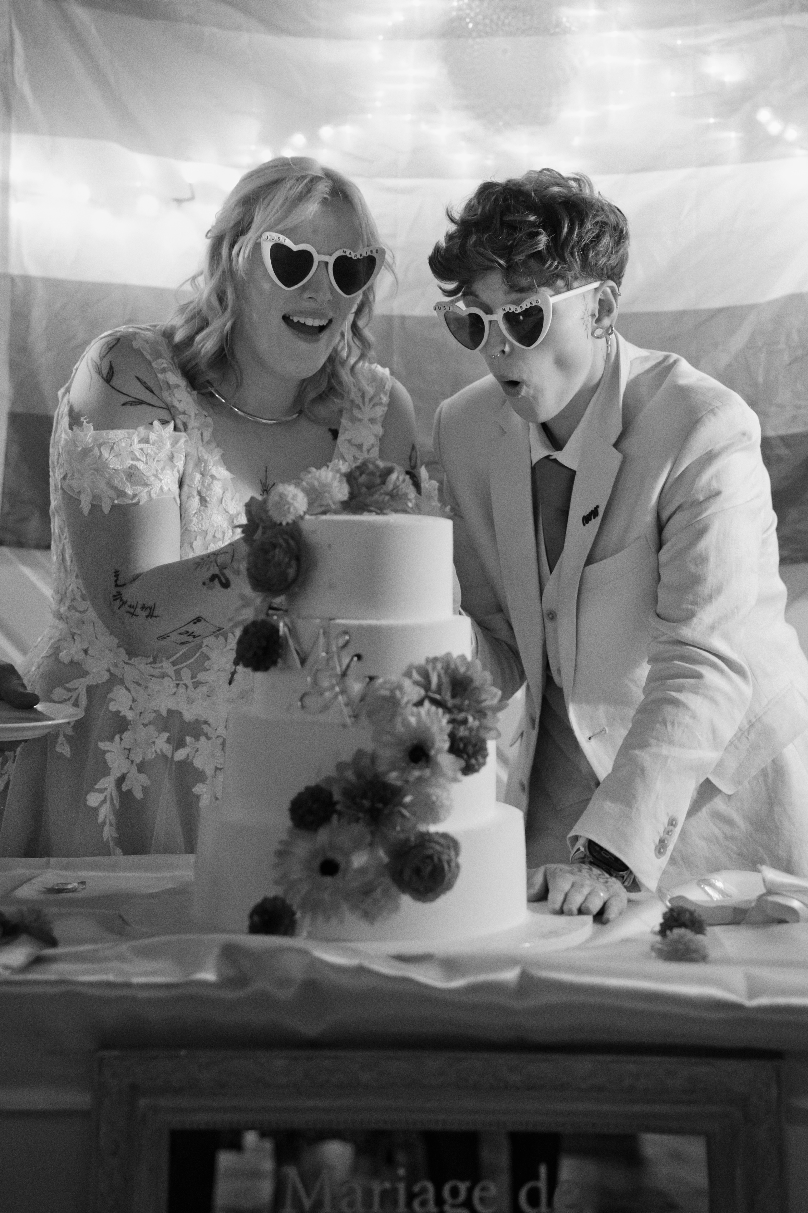 Deux personnes avec des lunettes en forme de cœur coupent un gâteau de mariage à étages décoré de fleurs.