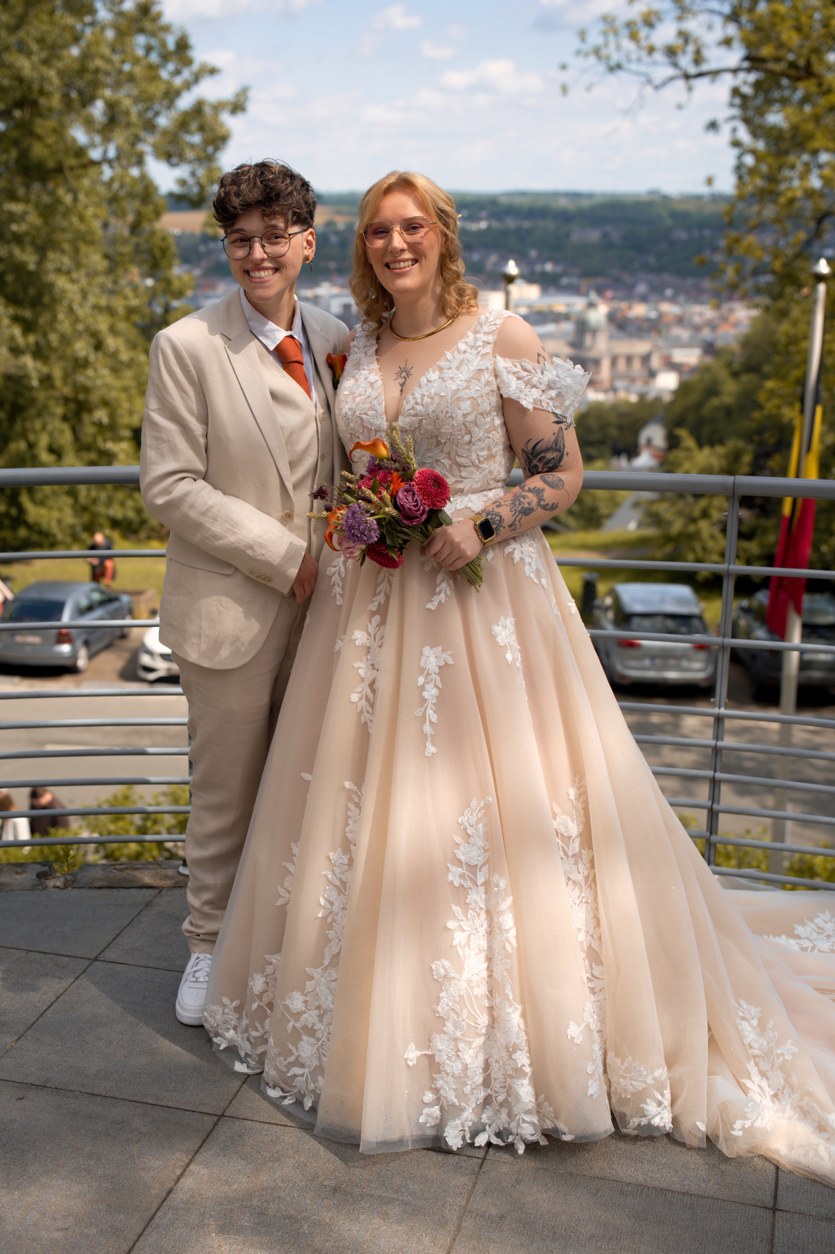 Couple mariée souriante, une en costume beige, l'autre en robe blanche fleurie tenant un bouquet.