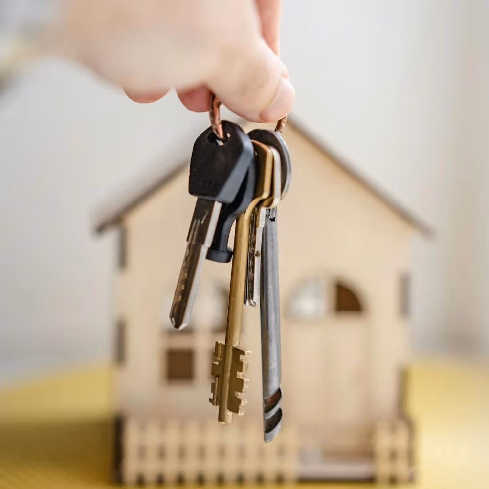 Hand holding a set of house keys with a wooden house in the background