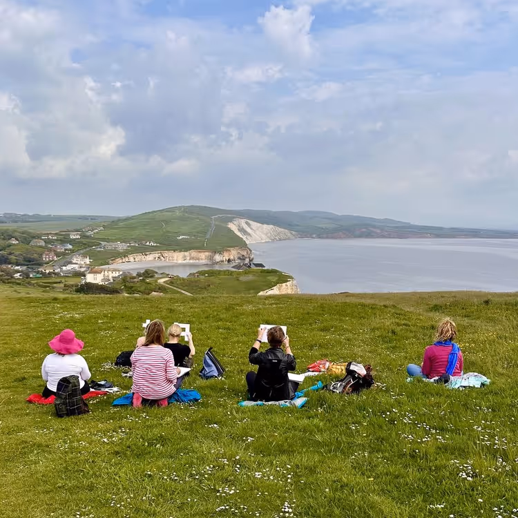 People sat on grass painting a coastal scene