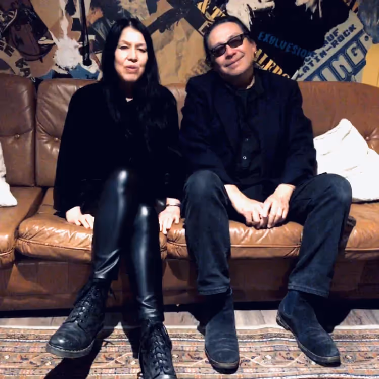 A man and woman dressed in black sitting on a brown leather sofa