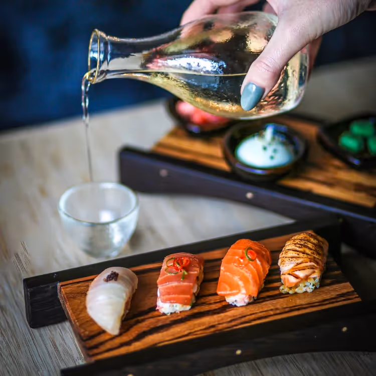 Sake being poured into a glass next to a dish of sushi