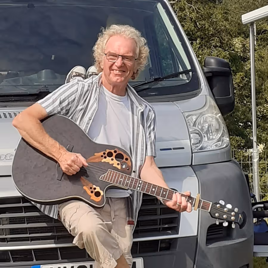 A man leaning against a van and holding a guitar