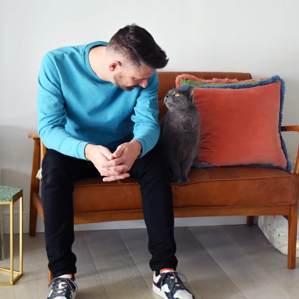 A man and a grey cat sitting on a small brown sofa.