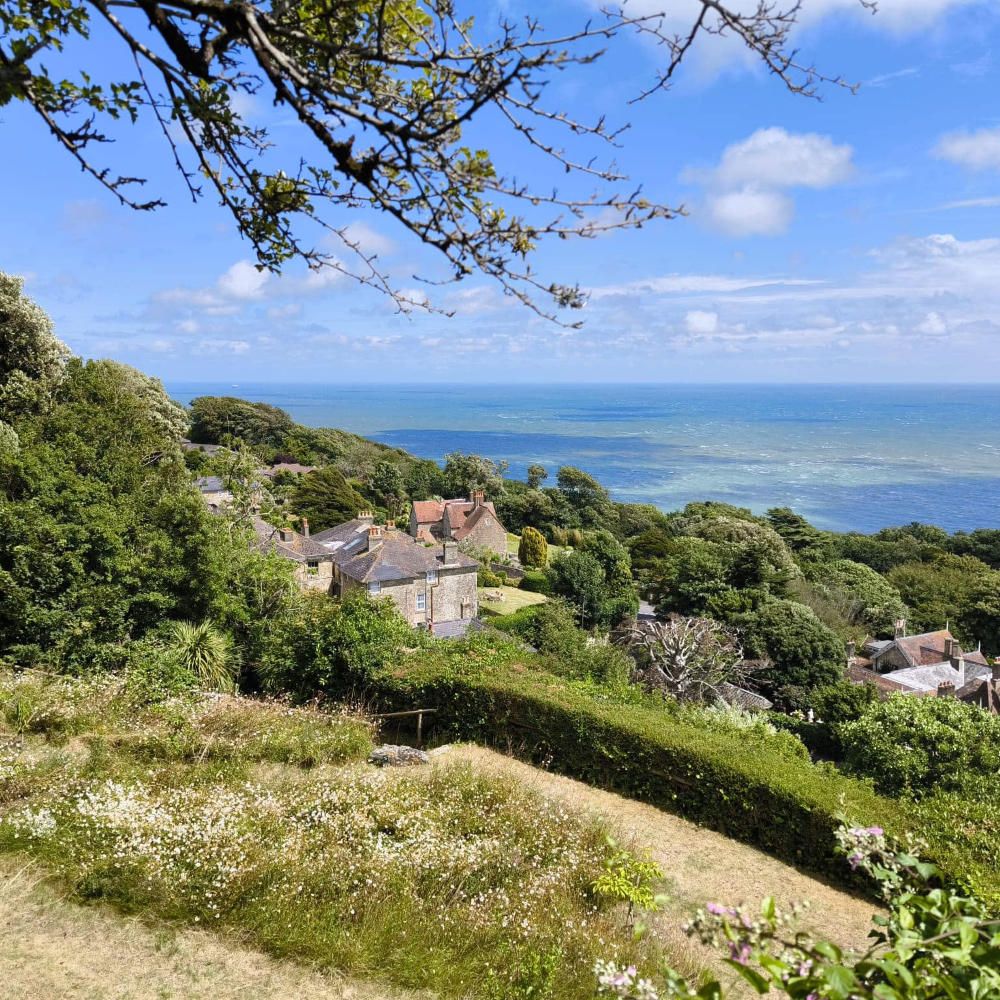 A view down a sloping hill to a stone house and the sea beyond.