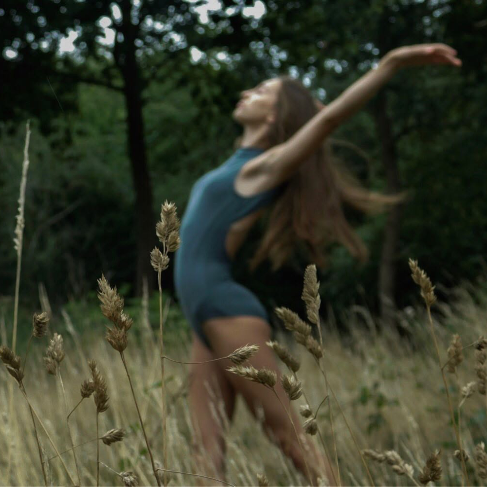A woman dancing amonst long grass beside woodland.