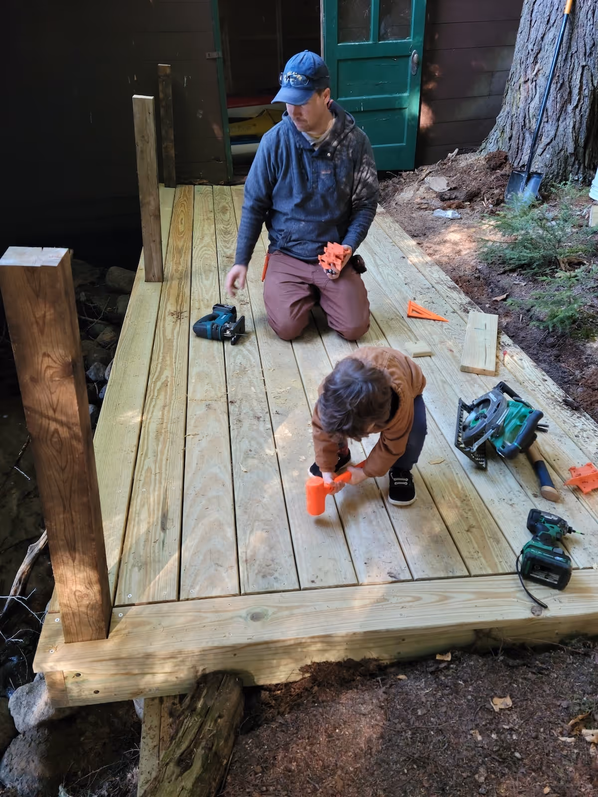 Man and child kneeling on a partially built wooden deck surrounded by tools, with the man holding orange shims and the child using a small orange hammer.