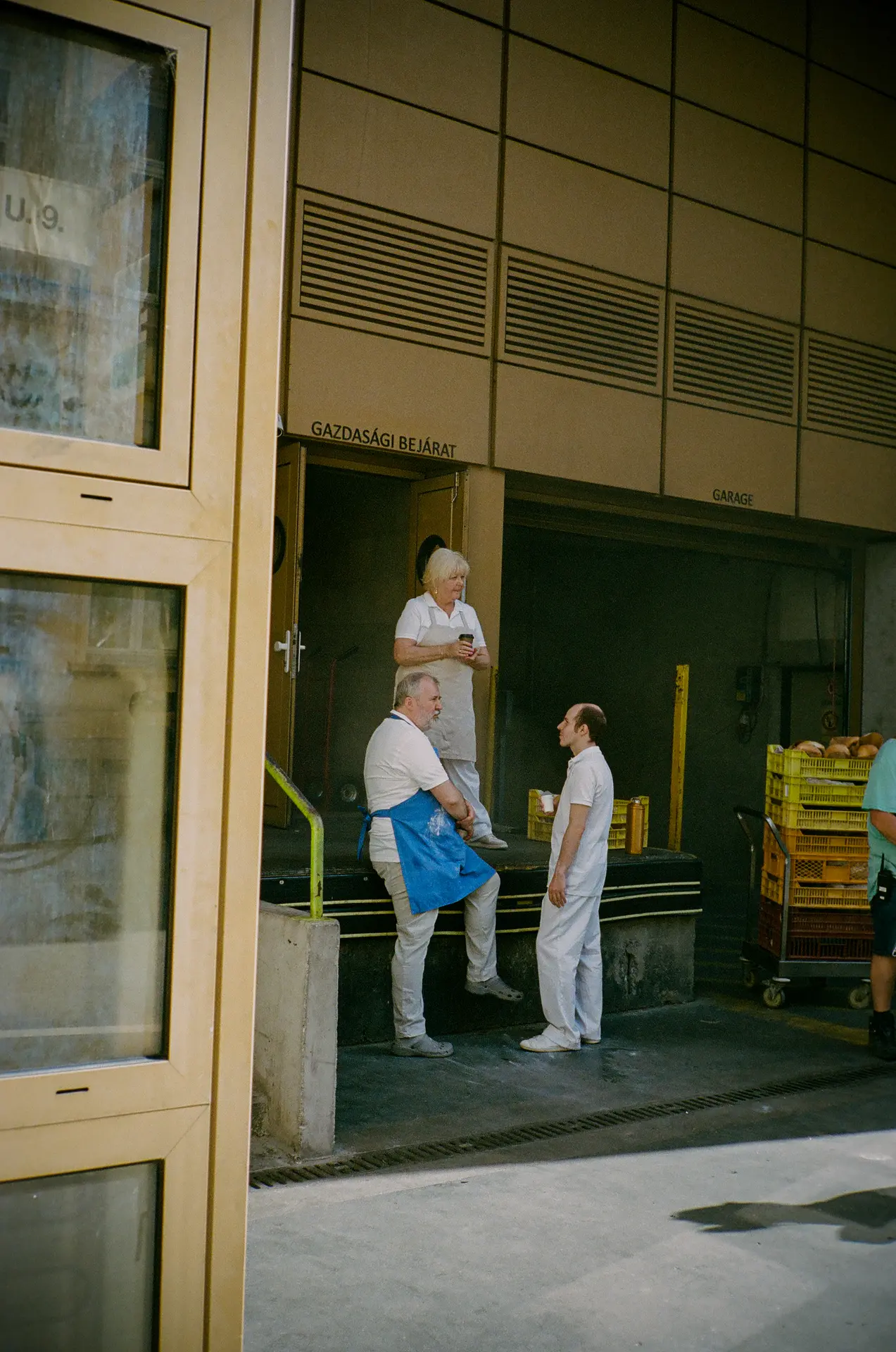 Three people in work uniforms chat and drink outside a building labeled "Gazdasági Bejárat," near stacks of yellow crates.