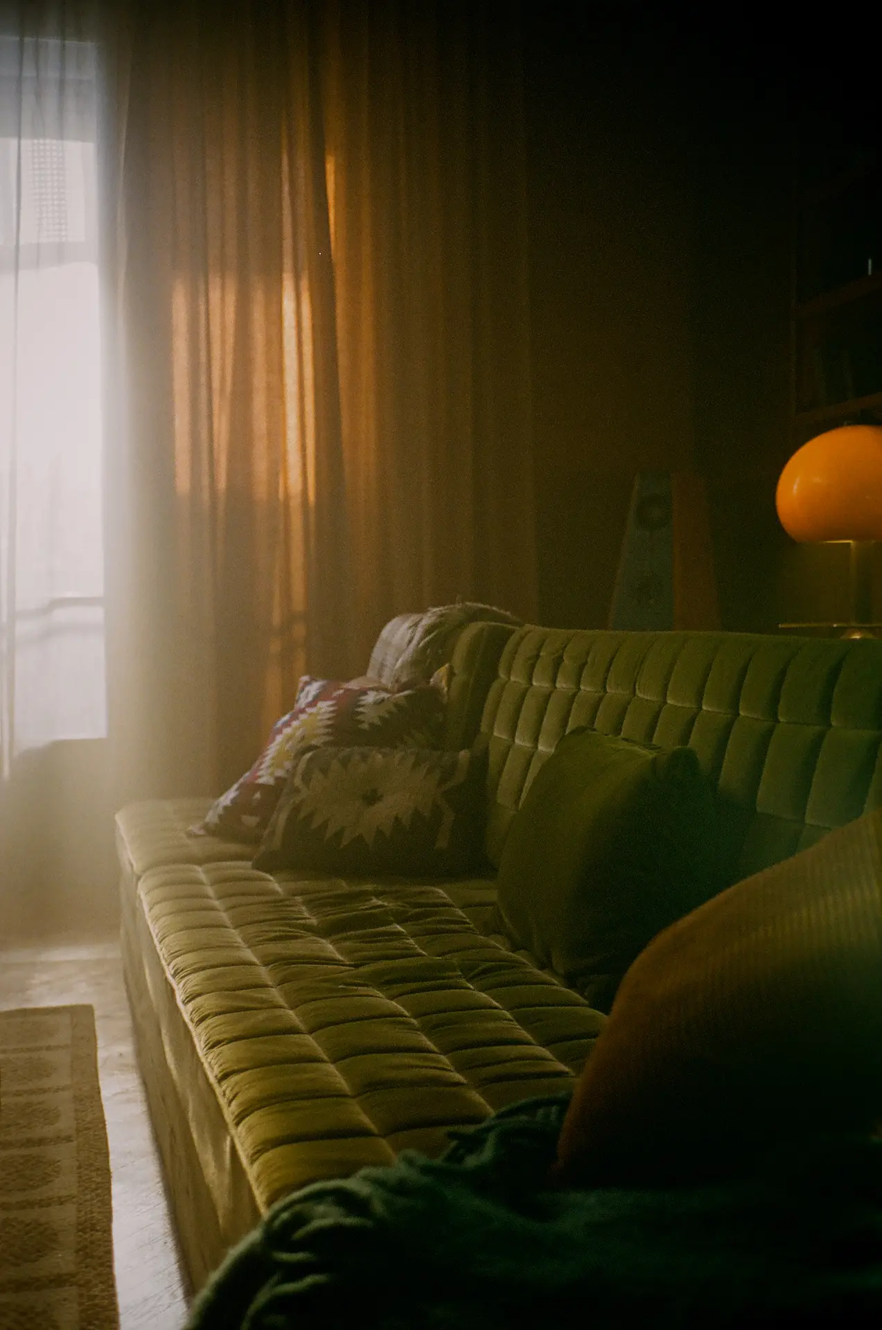 Cozy living room with a green couch and patterned cushions, warm light filtering through sheer curtains.