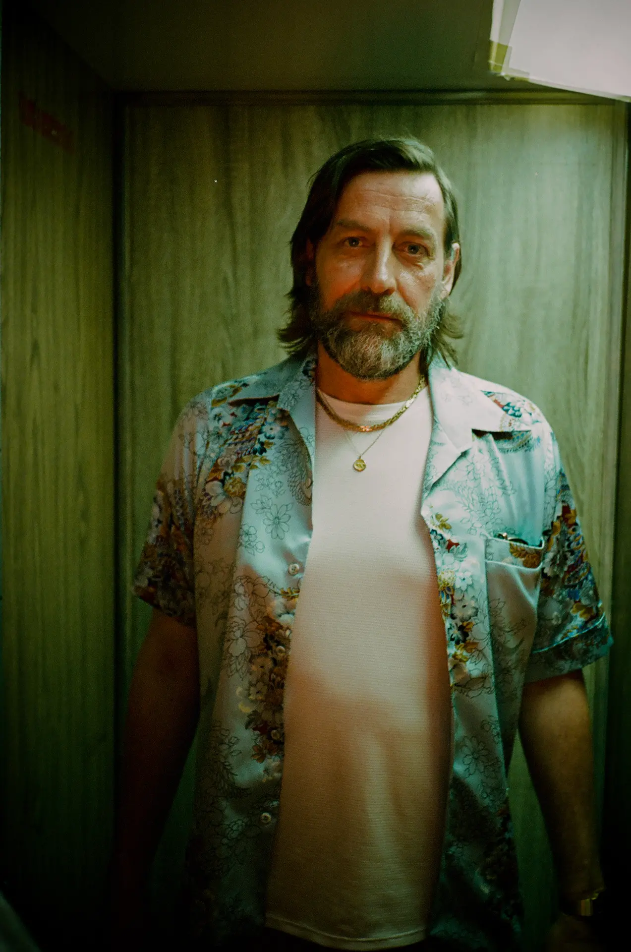 Portrait of a middle-aged man with a beard, wearing a floral shirt, standing in a dimly lit hallway.