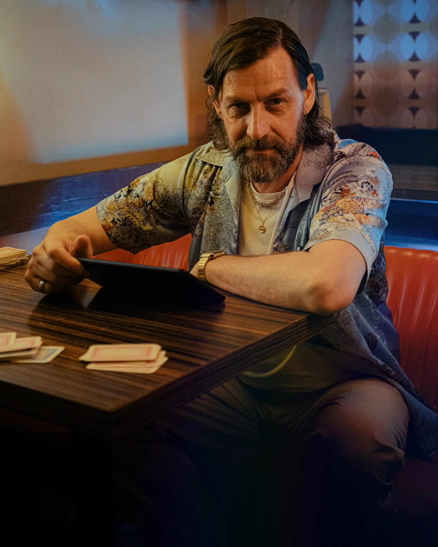 Middle-aged man with a contemplative expression, seated at a booth, looking at a tablet in a dimly lit diner.
