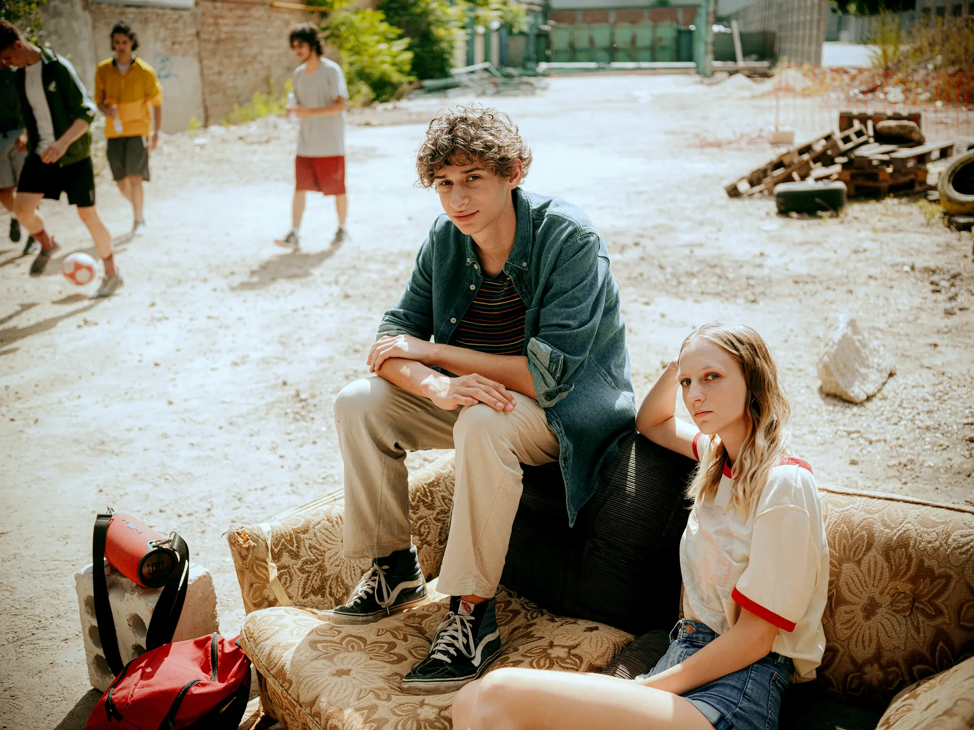 Young man and woman sitting thoughtfully on an old couch in a dilapidated urban yard while others play soccer nearby.