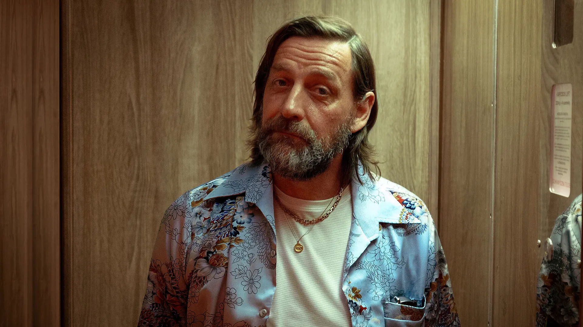 Middle-aged man with a beard in a floral shirt, standing inside an elevator with a reflective wood panel background.