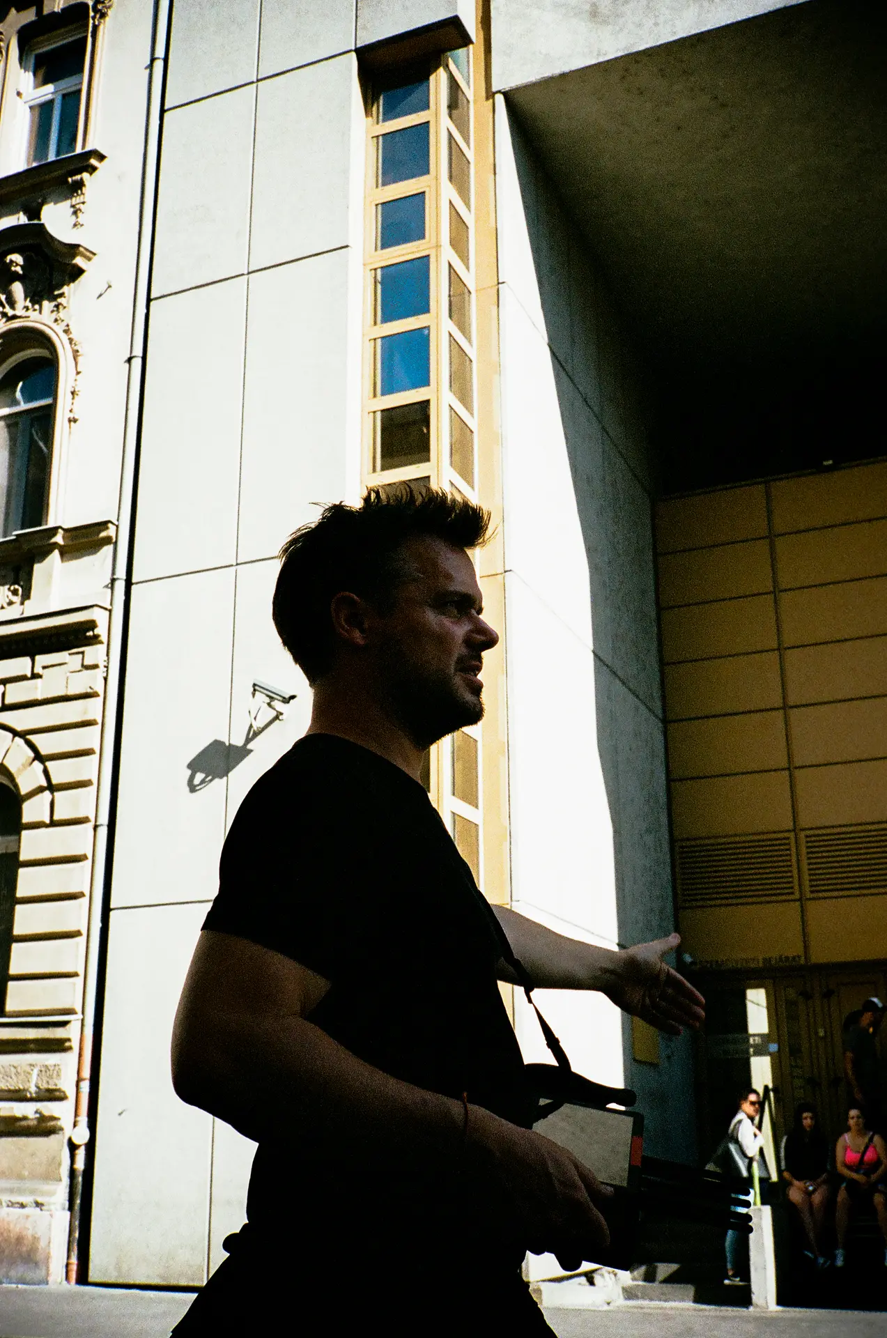 Young man with a confident stride walking past a building in a sunlit urban setting, casting sharp shadows.
