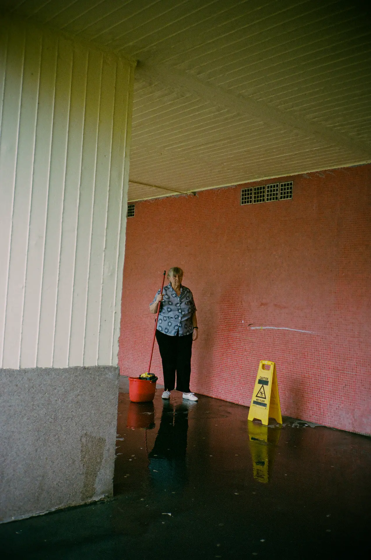 Elderly woman with a mop stands by a wet floor sign in a corridor with pink tiled walls and a reflective floor.