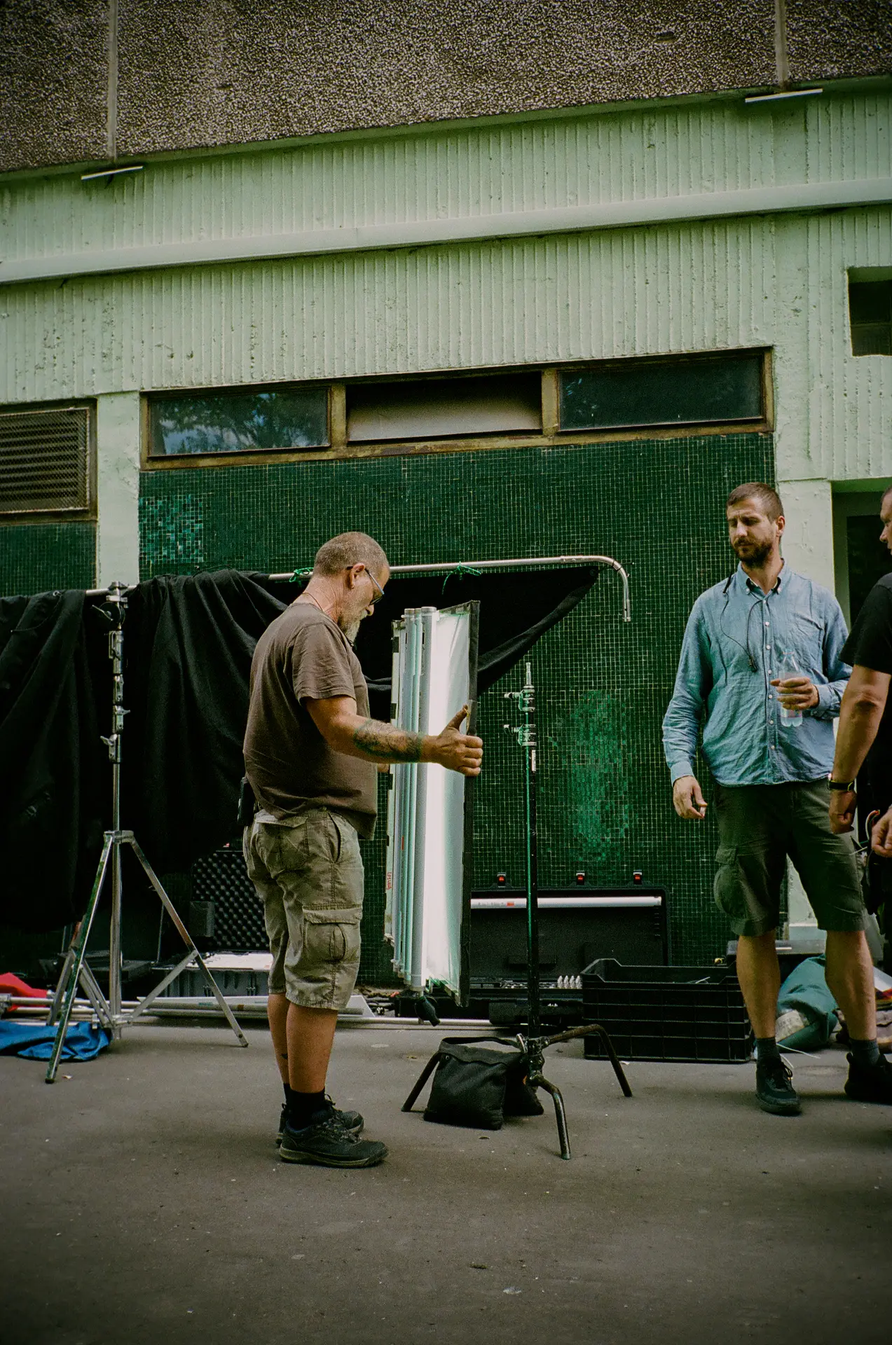 Man in cargo shorts setting up a light stand on a film set with another man observing.
