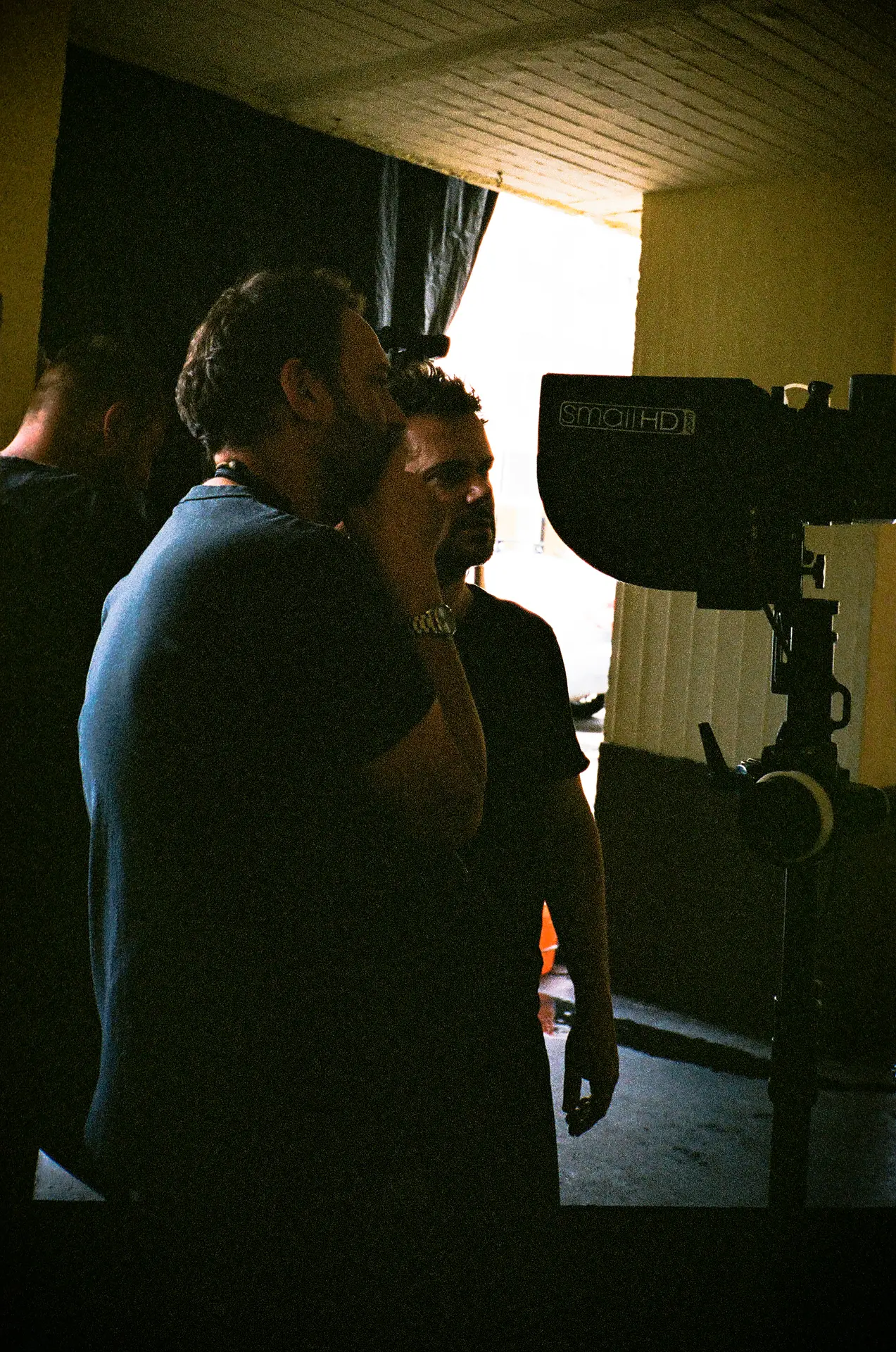 Two men, one touching his chin, discuss behind a camera on a film set, capturing a mood of focused collaboration.