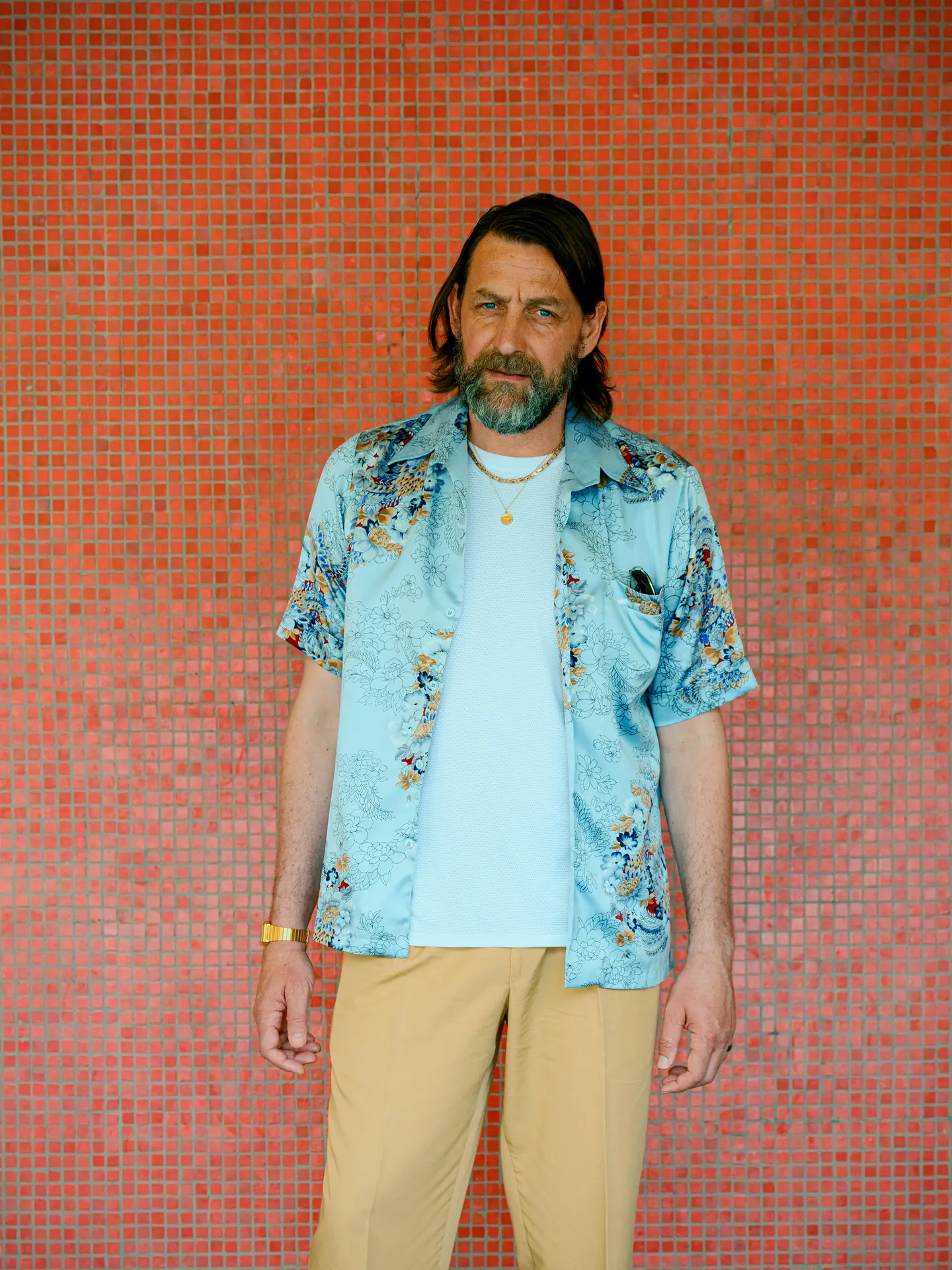 Man with a beard standing against a red mosaic wall, wearing a floral shirt and khaki pants, looking directly at the camera.