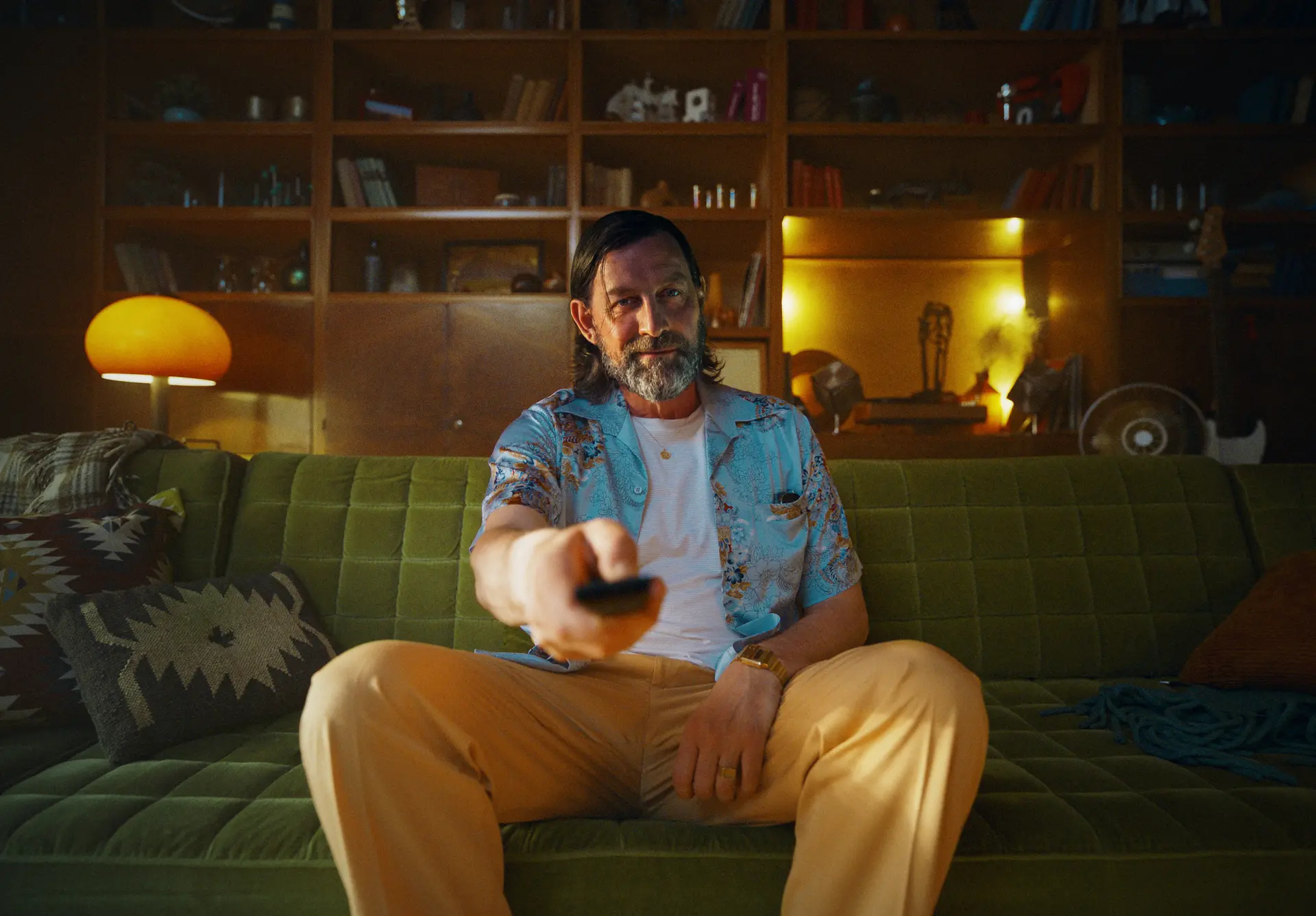 Middle-aged man with a beard, sitting on a green couch, extends a remote towards the camera in a cozy room.