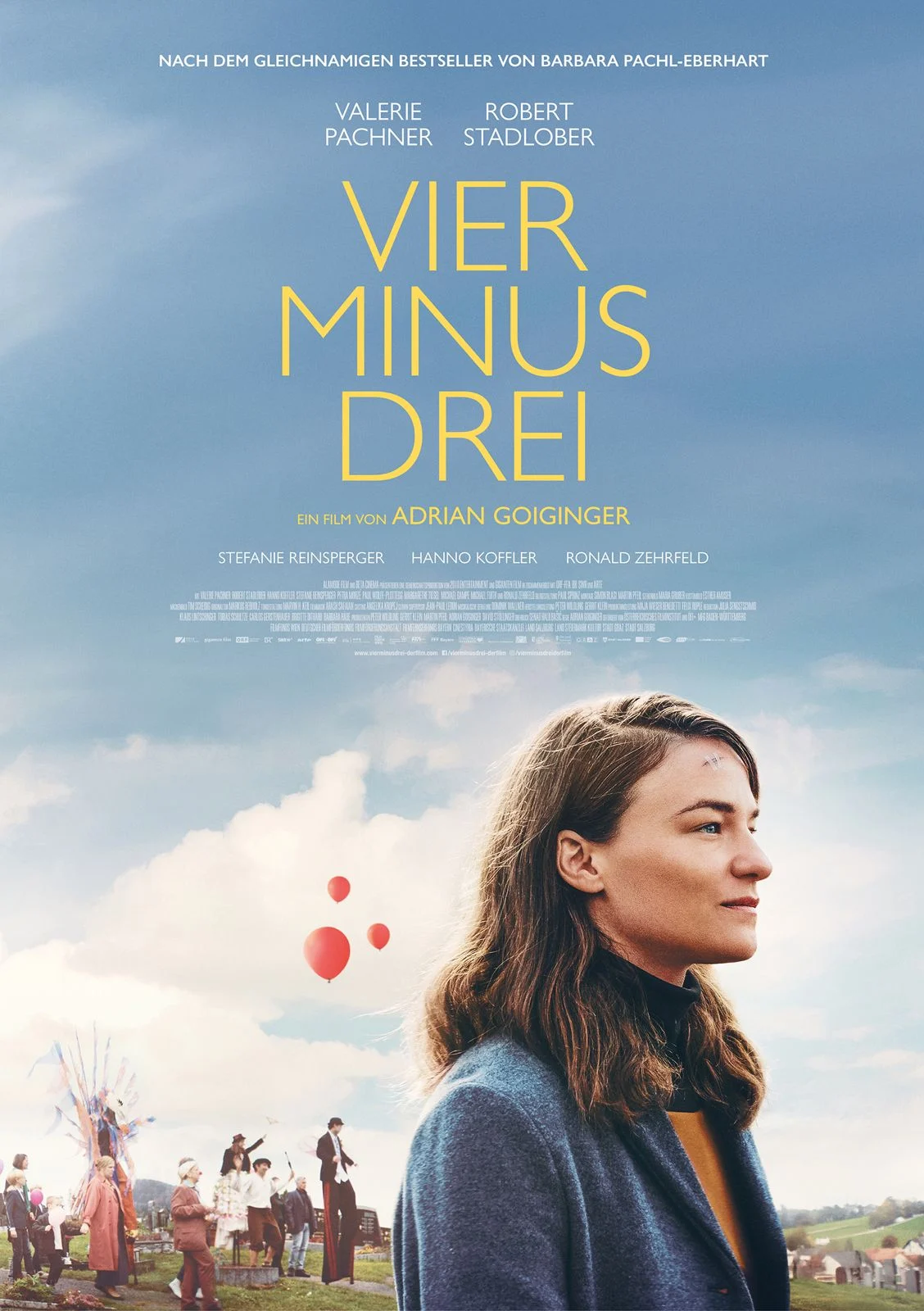 A movie poster showing a young woman in profile standing outdoors under a cloudy blue sky. She has shoulder-length brown hair and wears a blue coat and dark turtleneck. Behind her, a festive village scene unfolds with people gathered, colorful decorations, and several red balloons floating upward. In the sky above them, large yellow text reads “Vier Minus Drei.” Smaller text lists cast and crew details across the top and center of the poster.