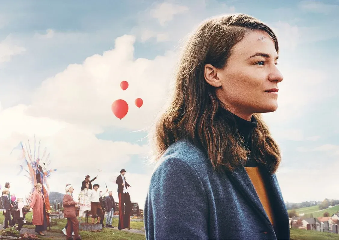 A movie poster showing a young woman in profile standing outdoors under a cloudy blue sky. She has shoulder-length brown hair and wears a blue coat and dark turtleneck. Behind her, a festive village scene unfolds with people gathered, colorful decorations, and several red balloons floating upward. In the sky above them, large yellow text reads “Vier Minus Drei.” Smaller text lists cast and crew details across the top and center of the poster.